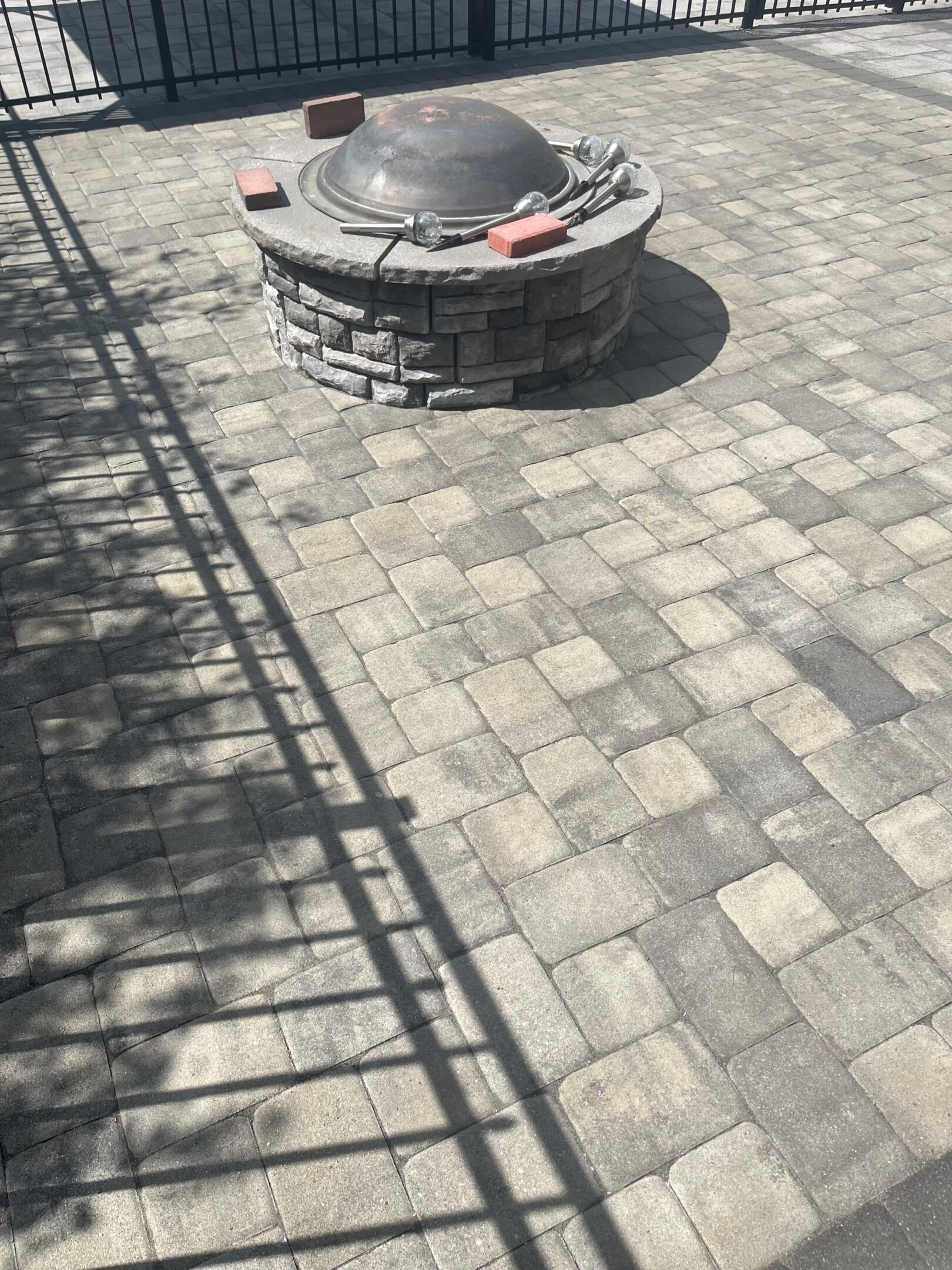 A fire pit on a paved stone patio with shadows from a nearby fence.